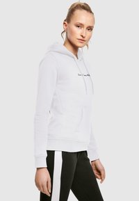 Biały hoodie z przednią kieszenią, kapturem ze sznurkiem i haftowanym czarnym napisem. W połączeniu z czarnymi spodniami z białymi paskami po bokach.