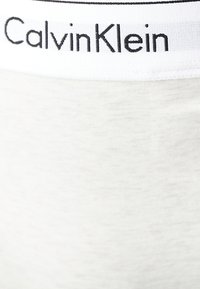 Calvin Klein midje i hvit, med svart logotekst. Stoffet nedenfor er en myk, lys grå nyanse med en jevn tekstur.