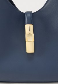 Tmavomodrá kožená kabelka s hladkou textúrou a zlatým zapínaním s jedinečným geometrickým dizajnom. Štruktúrovaný a elegantný tvar.