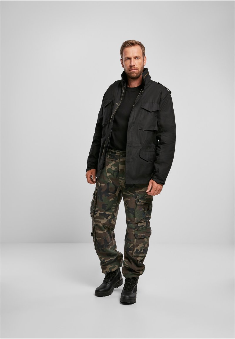 Schwarze Utility-Jacke mit zwei vorderen Taschen, kombiniert mit camosfarbenen Cargo-Hosen. Robuste schwarze Stiefel runden den Look ab, vor einem neutralen Hintergrund.