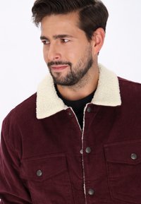 Bordeaux fløjlsjakke med cremesfarvet krave, to brystlommer og trykknapper. Blød tekstur og horisontal ribbet mønster.