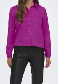Blusa fucsia de manga larga en una tela suave, con cuello y cierre de botones, combinada con pantalones negros ajustados.