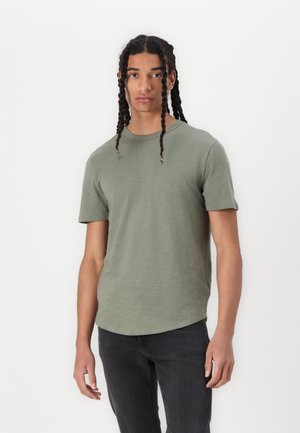 Olive green short-sleeve t-shirt, round neckline, soft texture, slightly curved hem, worn with black jeans.
