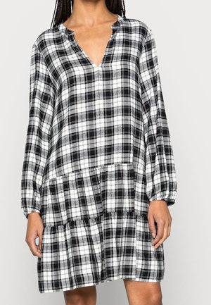 Czarna i biała sukienka w kratę z długim rękawem, dekoltem w serek i warstwową spódnicą, noszona przez stojącą osobę z warkoczami.