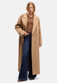 Płaszcz z mieszanki wełny wielbłądziej z kołnierzem z wycięciem, dużymi klapami i przednimi kieszeniami, zestawiony z brązową bluzką i ciemnymi jeansami dzwonami.