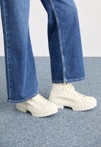 Sneaker alta bianche con suole spesse e rigate e dettaglio del logo circolare, indossate con jeans di denim azzurro chiaro a gamba larga.