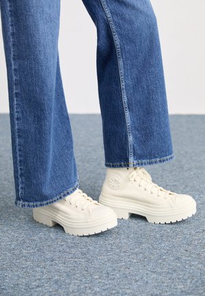 Ténis brancos de cano alto com solas grossas e estriadas e detalhe de logótipo circular, usados com jeans de ganga de perna larga em azul claro.