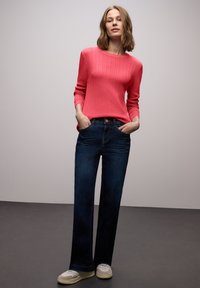 Czerwony prążkowany sweter z długim rękawem zestawiony z ciemnoniebieskimi jeansami o prostych nogawkach i jasnymi trampkami. Model stoi z rękami w kieszeniach.