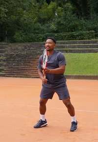 Tennis spiller iført en mørkegrå kortærmet skjorte og shorts, der holder en ketsjer, på en orange grusbane med grønne planter i baggrunden.