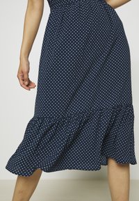 Žena v námořnické modré midi sukni s bílými puntíky a volánkovým lemem, stojící proti jednoduchému světlému pozadí.