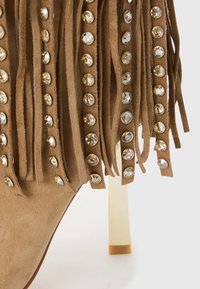 Tan suède hoge hak schoenen met franjes versierd met ronde gouden en zilveren rhinestones op een witte achtergrond.