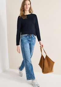 Femme portant un haut bleu marine à manches longues, un jean bleu et des baskets blanches, marchant en tenant un grand sac à main en daim marron.