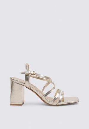 Sandale dorée métallique à brides avec talon bloc, bride de cheville à boucle et bout carré, vue de côté sur un fond uni.