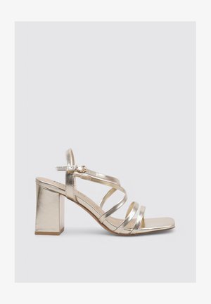 Sandale dorée métallique à brides avec talon bloc, bride de cheville à boucle et bout carré, vue de côté sur un fond uni.