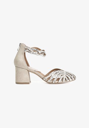 Beige suede heeled sandal with a block heel, featuring a crisscross strap adorned with metallic pyramid studs and an open-toe design.