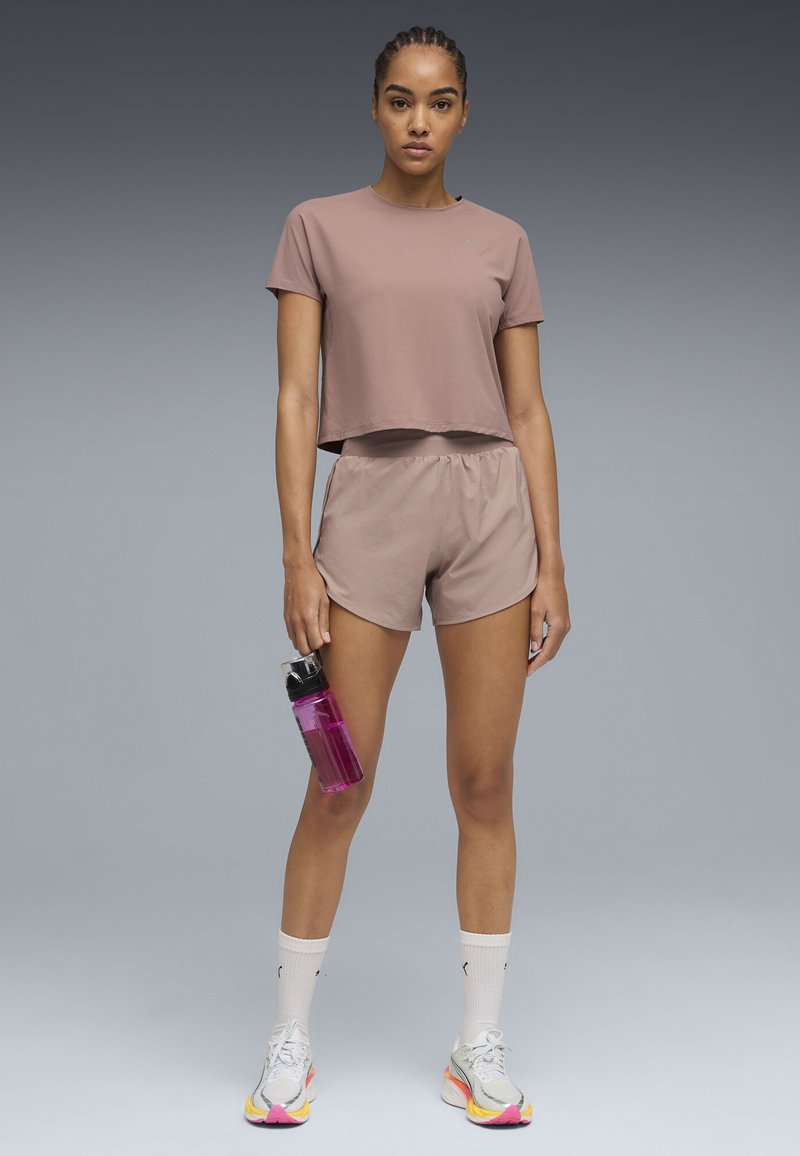 Γυναίκα που στέκεται φορώντας μοβ αθλητικά σορτς και κορμάκι, κρατώντας ένα ροζ παγούρι νερού, με λευκές κάλτσες και πολύχρωμα αθλητικά παπούτσια.