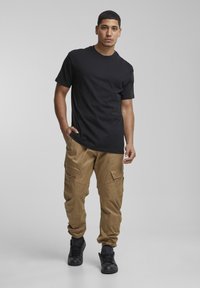 Črna bombažna majica v kombinaciji s kaki cargo hlačami z več žepi, sproščenim krojem in črnimi supergami. Enostavno ozadje.