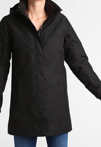 Žena oblečená v dlouhé černé bundě na zip s límcem, doplněná modrými džíny, stojící na pozadí s jednobarevným světlým pozadím.