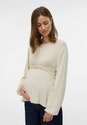 Ljusbeige mammatröja i mjukt tyg, med långa ärmar, en draperad midja och en avslappnad siluett. Parat med mörk denim.