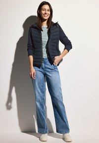 Giacca trapuntata blu navy con cappuccio e chiusura con zip, abbinata a una maglietta a righe blu chiaro e a un paio di jeans a gamba larga blu chiaro. Le sneakers bianche completano il look.