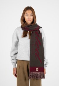 Mørkebrun og burgunder tørklæde med logo-design, frynser i enderne og en glat tekstur, båret over en lys grå sweatshirt.