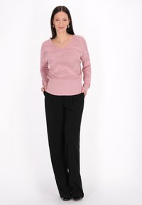 Pulover tricotat roz deschis cu decolteu în V, având un model texturat, asortat cu pantaloni lejeri negri. Modelul pozează cu mâinile în buzunare.