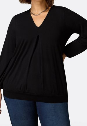 Kvinde iført en sort langærmet V-hals bluse med en centerforan-plissé og mørkeblå jeans, poserer med den ene hånd på hoften.