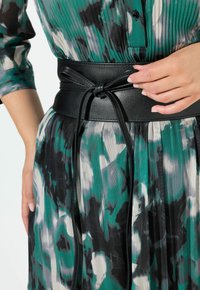 Cintura in pelle nera con un dettaglio annodato, indossata sopra un abito plissettato con pattern verde, nero e bianco. Texture liscia e design su misura.
