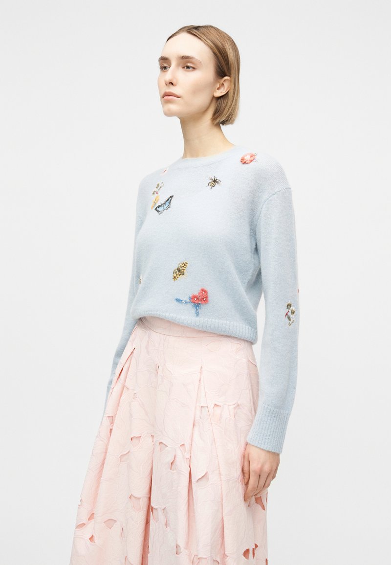 Donna che indossa un maglione azzurro chiaro con farfalle e fiori ricamati, abbinato a una gonna rosa pallido con texture.