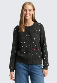 Lächelnde Frau in einem dunkelgrauen, geknöpften Cardigan mit weißen und roten Herz- und Punktmustern, kombiniert mit blauen Jeans, stehend vor einem hellen Hintergrund.