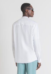 Camicia bianca a maniche lunghe con una superficie liscia, polsini con bottoni e orlo curvo, abbinata a pantaloni azzurri chiari, vista da dietro.