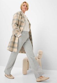 Rutig uldfrakke i beige og grå, hvid bluse, grå stribede bukser og lyse loafers, poseret selvsikkert mod en neutral baggrund.