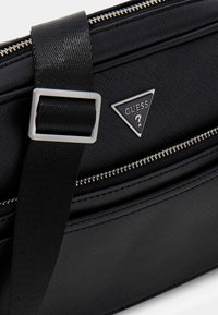 Sort stof tas med tekstureret overflade, sølv lynlås og justerbar strap. Har et trekantet sølv logo på fronten.