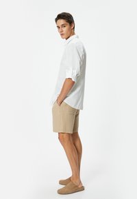 Camicia bianca in lino con maniche arrotolate, pantaloni corti beige testurizzati e scarpe slip-on marroni, in profilo contro uno sfondo bianco uniforme.