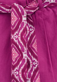 Roza tkanina zložena s kravato, ki ima vzorec geometrijskih oblik v belem in temno roza. Gladka tekstura, sproščen dizajn in vidne šive.