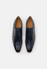 Scarpe eleganti in pelle blu scuro con dettagli brogue, chiusura con lacci e finitura liscia. Presentano una punta arrotondata e una suola color beige chiaro.