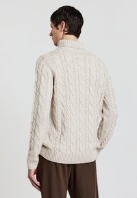 Beiger Kabelstrickpullover mit hohem Kragen, gerippten Bündchen und Saum. Verfügt über ein strukturiertes Muster und eine lockere Passform. Getragen mit braunen Hosen.