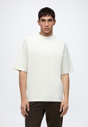 Jeune homme aux cheveux courts et foncés portant une chemise blanche unie oversize à manches courtes et un pantalon foncé, debout sur un fond blanc.