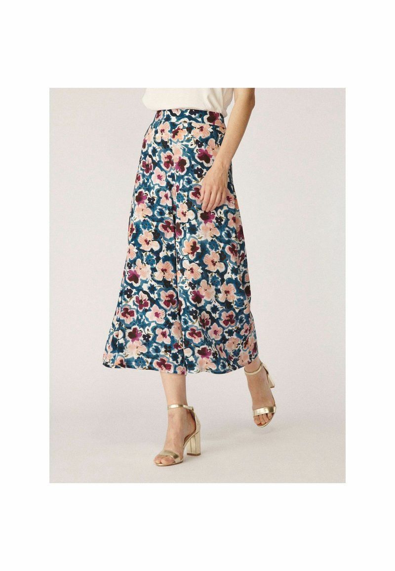 Femme portant une jupe taille haute mi-longue à imprimé floral bleu, rose et bordeaux, associée à des sandales à talons blocs dorés.