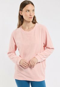 Chemise rose à manches longues avec une texture côtelée, un col rond et des détails de couture visibles. Portée avec un bas bleu.