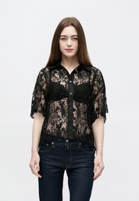 Sort blondebluse med korte ærmer og knapper foran; blomsterprint; ensfarvet sort krave og knapper; båret med mørke denimbukser.