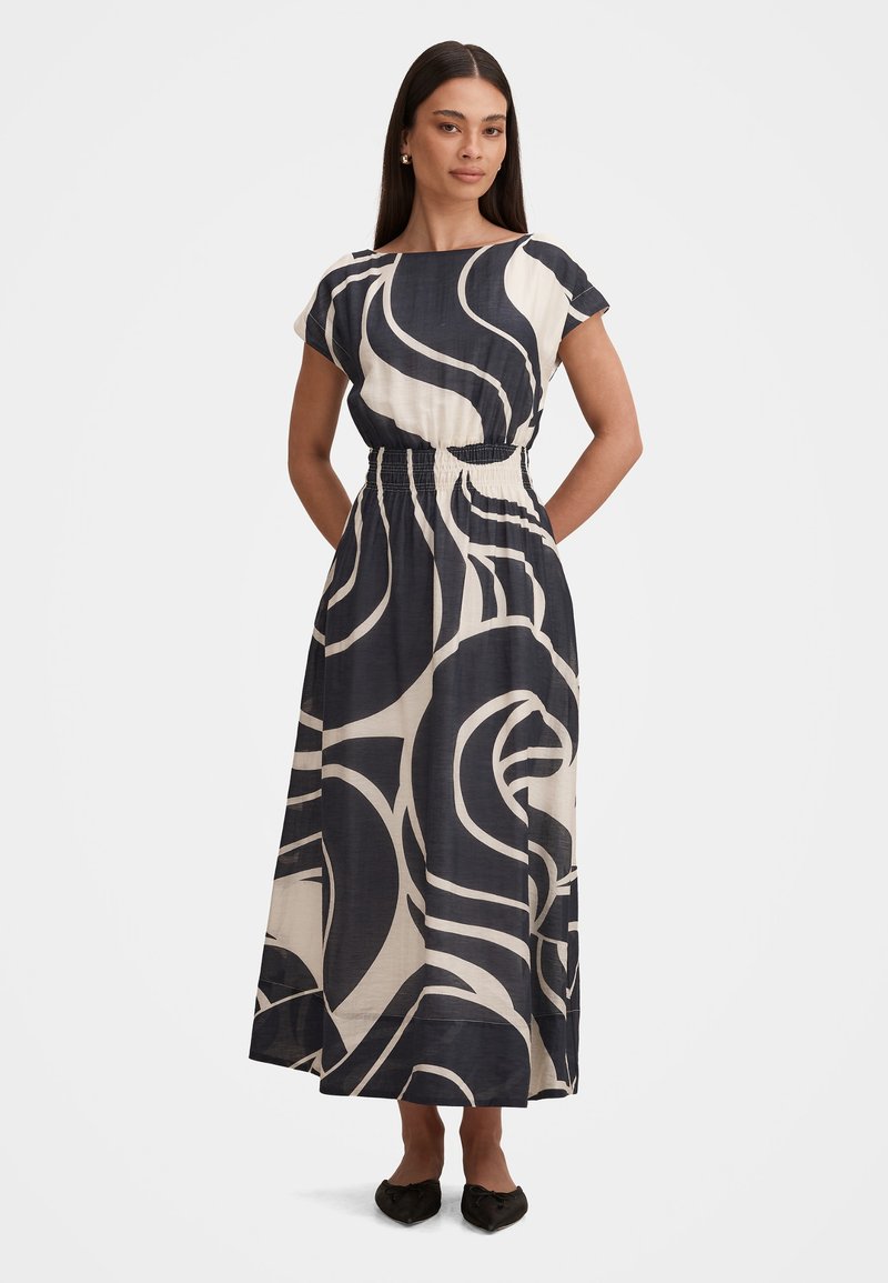 Γυναίκα που φοράει midi φόρεμα με μαύρο και λευκό αφηρημένο σχέδιο, κοντά μανίκια, ελαστική μέση και μαύρα μυτερά παπούτσια, στέκεται με τα χέρια πίσω.