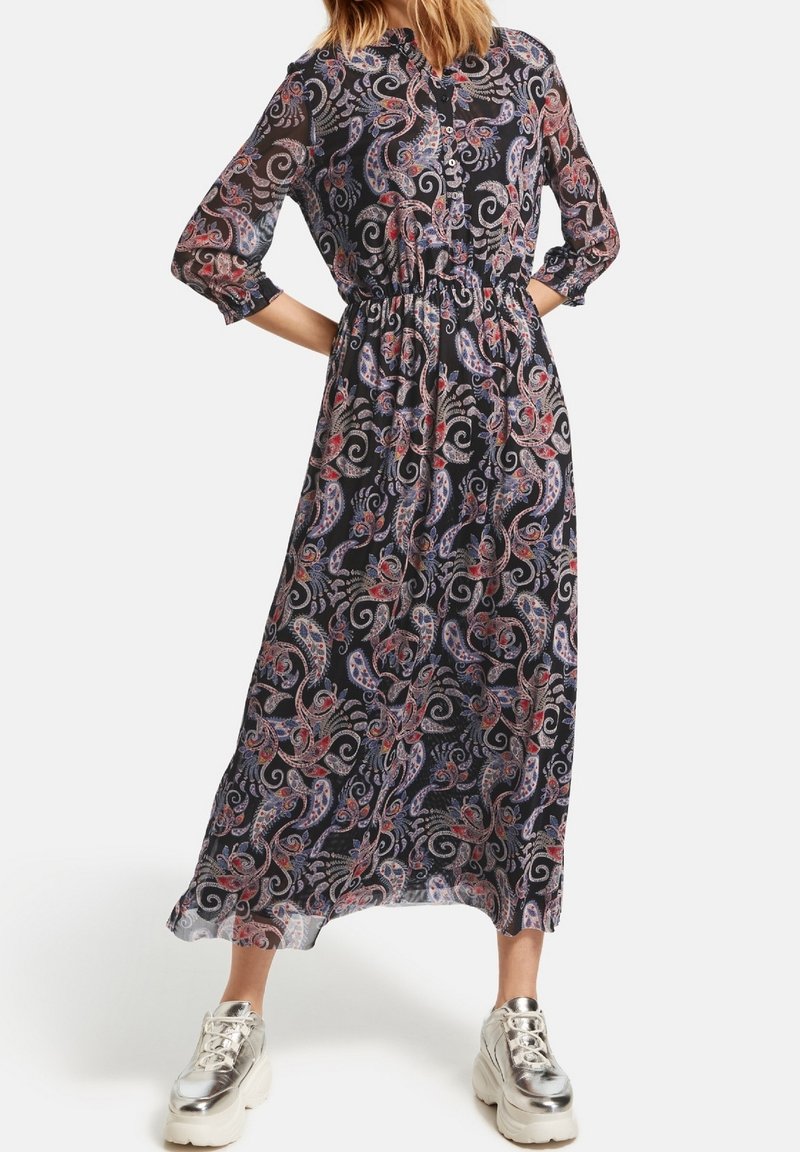 Robe longue et transparente noire avec un motif paisley en bleu, rouge et blanc, dotée d'un devant à boutons et d'une taille élastique, associée à des baskets argentées.