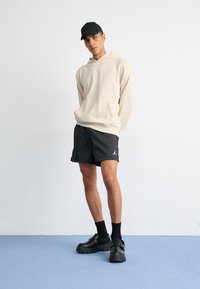 Beige hoodie with kangaroo pocket, black athletic shorts with logo, and chunky black shoes. Model wears a black cap. Light background.