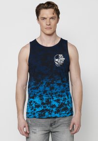 Modra gradientna majica brez rokavov z grafikonom lobanje in besedilom; izdelana iz bombaža z mehkobnim, teksturiranim zaključkom. Model nosi sive oblečene jeans hlače.