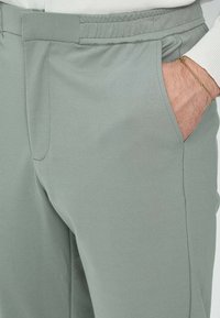 Pantalon léger vert clair à taille élastique, fabriqué à partir d'un tissu lisse et léger. Dispose d'une poche latérale et d'une coupe ajustée avec de subtils détails de plissage.