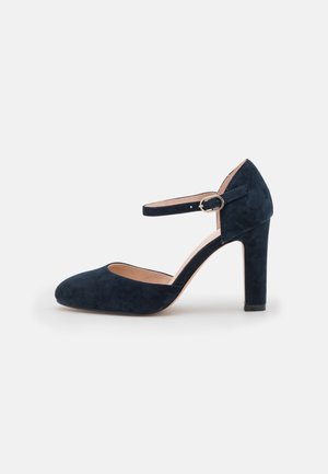 Chaussure à talon haut en daim noir avec bride de cheville et boucle dorée, bout fermé arrondi, talon bloc moyen sur fond blanc.
