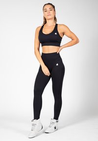 Black sports bra and high-waisted leggings in smooth fabric, featuring a small logo. Footwear is white with black accents and textured materials.