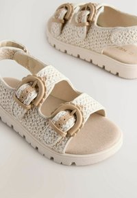 Weiße geflochtene Sandalen mit zwei verstellbaren, hellbraunen Riemen mit runden Schnallen, einer weichen beigen Fußbett und einer strukturierten Gummisohle.