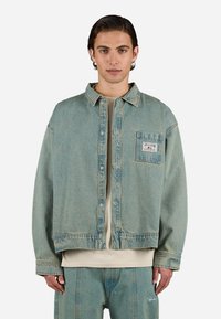 Chaqueta de denim azul claro con forma cuadrada, bolsillo frontal y cierre de botones. Presenta una textura desgastada y detalles de costura visibles.